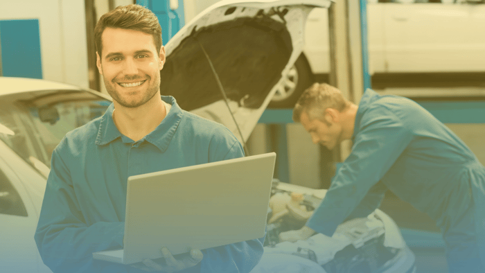 technician holding a laptop with another technician working under the hood of a car in the background