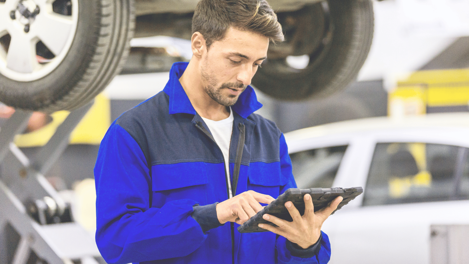 Technician in a blue jacket standing in front of a car on a lift looking at a tablet