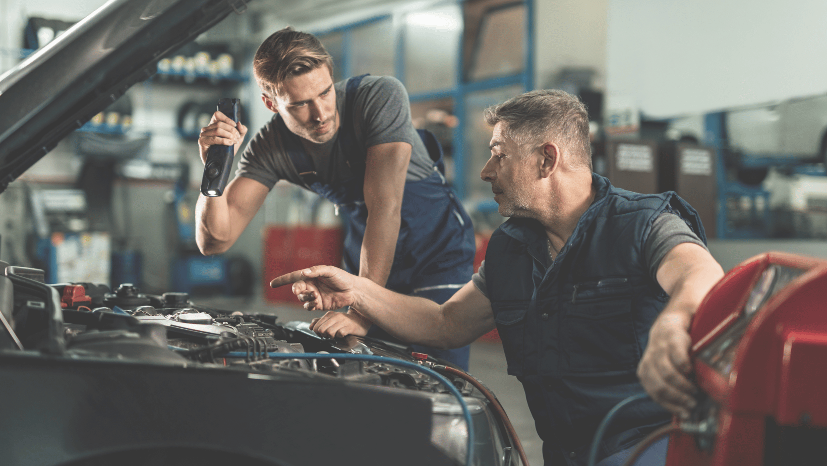 one technician charging a car battery with another technician holding a flashlight